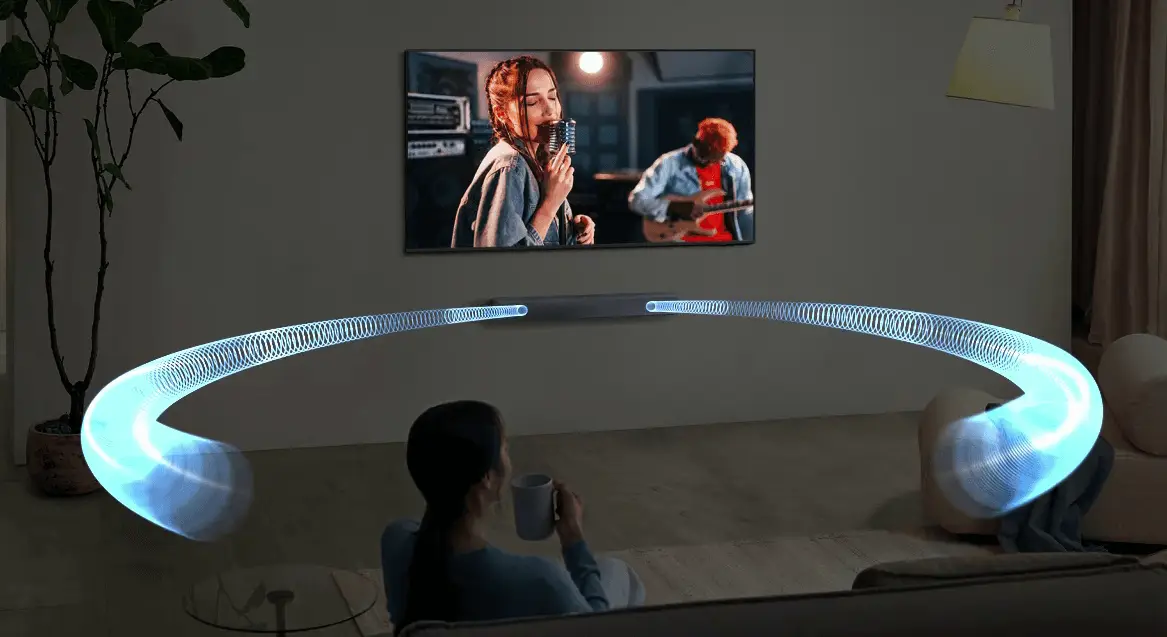 Uma pessoa sentada no sofá em uma sala escura, assistindo TV com uma soundbar Samsung abaixo da tela; ondas sonoras gráficas em azul saem do aparelho, ilustrando a dispersão do som pelo ambiente.