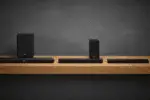 Sistema home theater JBL Bar: soundbar preta longa em mesa de madeira, subwoofer cúbico à esquerda, dois alto-falantes surround à direita, fundo escuro estúdio.